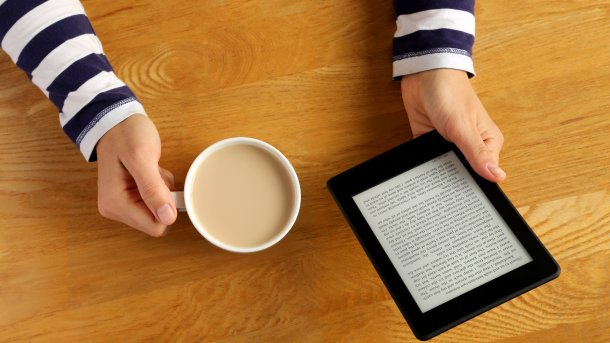 Eine Frau liest auf einem E-Reader ein Buch und trinkt dabei einen Kaffee.