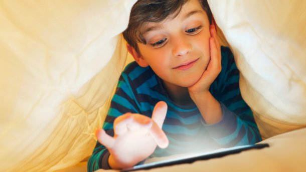 Junge tippt unter einer Bettdecke lächelnd am Smartphone
