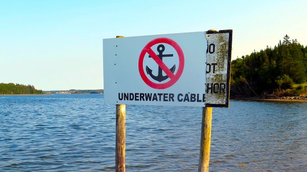 Ein Schild steht auf Holzbeinen im Wasser; es verbietet ankern weil dort ein "underwater cable" verläuft; im Hintergrund grüne Inseln