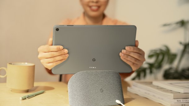 Ein mit grauem Textil bespannter Lautsprecher steht auf einem Tisch. Hinter dem Lautsprecher hält eine Frau die Rückseite eines grauen Tablets in die Kamera.