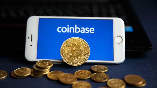 Ein Smartphone zeigt den Schriftzug "coinbase", davor liegen güldene Münzen; eine davon zeigt das Bitcoin-Logo