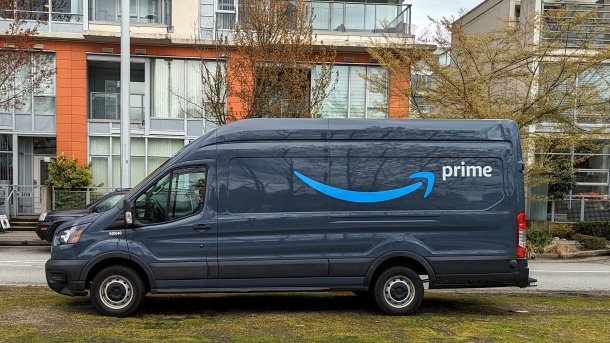 Ein dunkelgrauer Amazon-Lieferwagen mit Aufdruck "Prime" steht auf einem Grasstreifen, dahinter Wohngebäude