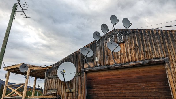 Holzgebäude, an dem 8 Satellitenschüsseln angebracht sind; links ein Mast mit einer terrestrischen Antenne