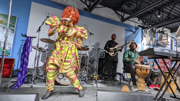 Schwarze Frau in gelbem Kleid tanzt auf Bühne und singt; rechts dahinter ein schwarzer Mann trommelt, ein weitere spielt Bass