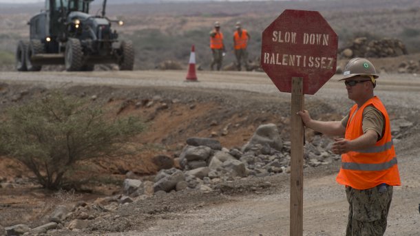 Soldat in Warnweste mit Stoptafel mit Aufschrift "Slow Down - Ralentissez"
