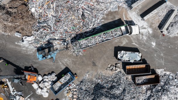 Luftaufnahme eines Aluminium-Recyclingbetriebs mit Metallschrotthalden, Schüttmulden, Lastwagen und einem Ladebagger.
