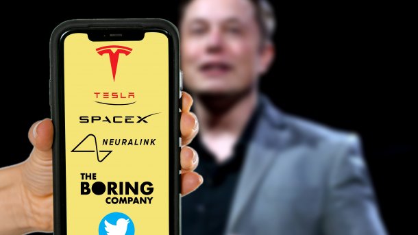 Ein Smartphone zeigt Logos von Tesla, SpaceX, Neuralink, The Boring Company und Twitter; hinter dem Handy ist Elon Musk zu sehen