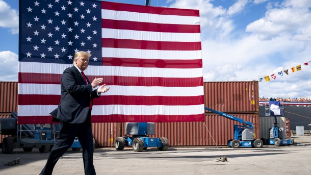 Donald Trump gehend und klatschend; im Hintergrund eine enorme US-Fahne, dahinter rostbraune Container