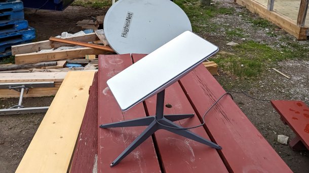 Starlink-Schüssel steht auf einem Holztisch; im Hintergrund eine demontierte Satellitenschüssel mit Aufschrift "HughesNet"
