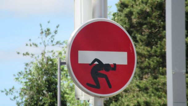 Verkehrszeichen "Ausfahrt einer Einbahnstraße" mit zusätzlich aufgemalter Figur, die quasi den weißen Querbalken auf der Schulter schleppt