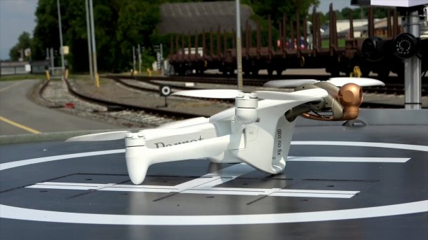 Weißer Quadkopter steht einer grauen Plattform, auf der ein großes weißes H in einem weißen Kreis aufgemalt ist; im Hintergrund Bahngeleise