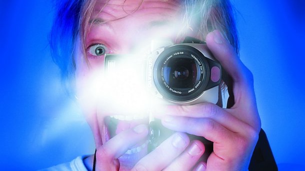Foto tersebut memperlihatkan kepala seorang pria dengan latar belakang biru, memotret dirinya sendiri dengan flash, satu mata terlihat, dan mata lainnya melihat melalui lensa. 