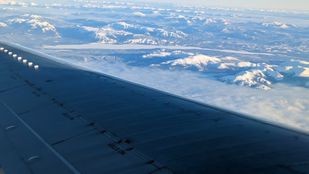Foto aus einem Flugzeug; im Vordergrund eine Tragfläche, unten verschneite Gebirge
