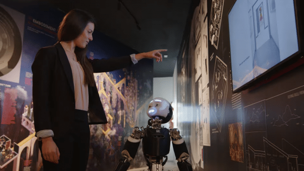 Eine Frau zeigt auf eine Leinwand; dazwischen steht ein kleiner Roboter, der die Frau anblickt