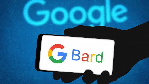 Eine Hand hält ein Handy, auf dessen Bildschirm das Google-Logo "G" sowie der Schriftzug Bard zu sehen sind; im Hintergrund leuchtet das Wort Google