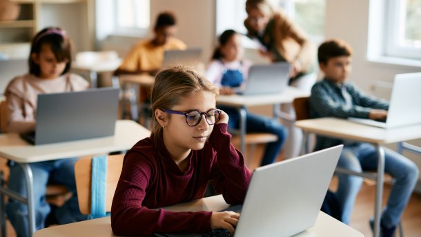 Eine Grundschülerin mit einer Brille sitzt mit anderen Kindern in einem Klassenzimmer und liest etwas auf einem Laptop.