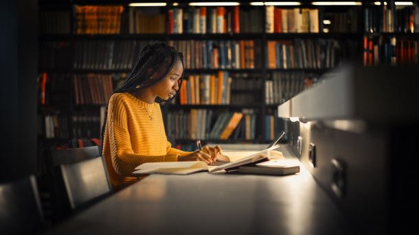 Mensch vor einem Bücherregal am Schreiben einer "längerfristigen" Arbeit, wahrscheinlich in einer Universitätsbibliothek