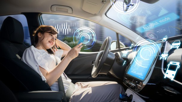 Lächelnde junge Frau am Volant eines fahrenden, vernetzten Autos im Autopilot-Modus; sie beschäftigt sich mit ihrem Handy