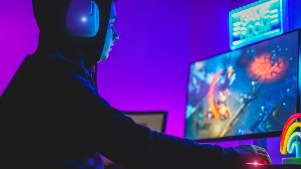Junger Gamer mit Hoodie und Kopfhörern vor einem Computerbildschirm. Er spielt in einem abgedunkelten Raum mit lila Beleuchtung ein Videospiel.