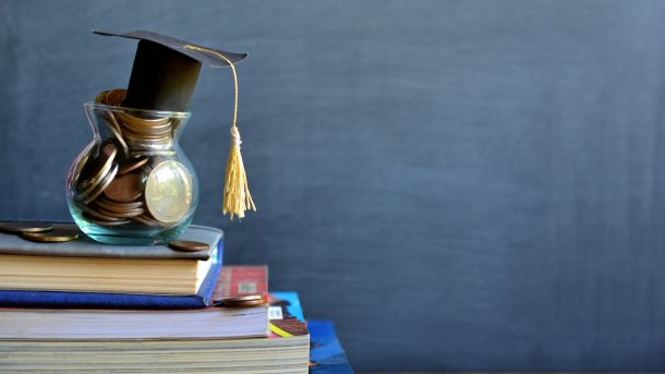 Ein kleines Glas mit Münzen, auf dem ein aus Pappe nachgebildeter Graduierten-Hut liegt, auf einem Stapel Schulbücher. Im Hintergrund eine Schultafel.