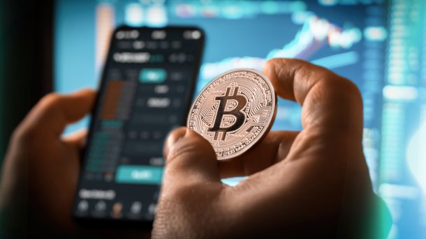 Hand mit Bitcoin vor Trading-App
