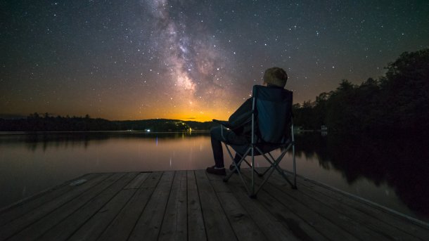 Mann sitzt auf Campingstuhl auf einem Steg unter dem Sternenhimmel