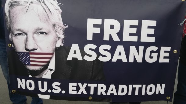 Transparent "FREE ASSANGE - no US extradition" mit Bild Julian Assanges; seinen Mund überklebt eine US-Fahne