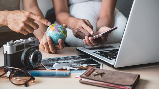 Zwei Menschen sitzen vor dem Laptop. Einer hält eine Weltkugel in der Hand, der andere ein Smartphone. Kamera, Kalender und Co. liegen auf dem Tisch