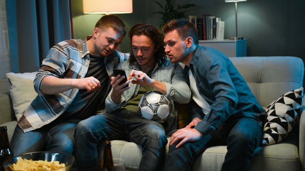Drei Männer sitzen auf dem Sofa und starren auf ein Handy, der mittlere Mann hat einen Fußball unter den Arm geklemmt.