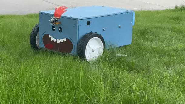 Man sieht den Mähroboter in Blau auf einer Wiese. Der Mäher hat ein aufgemaltes, breites Grinsen.