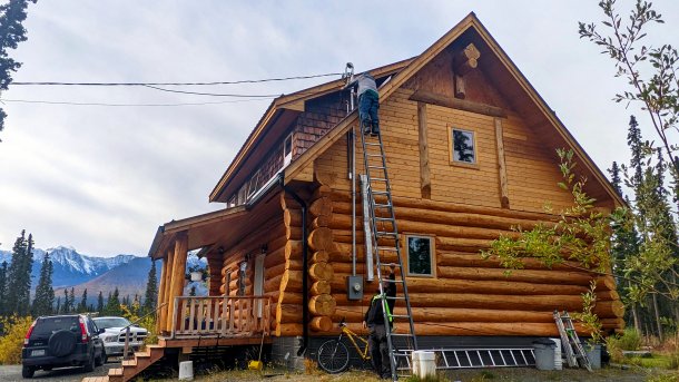 Hozhaus; Arbeiter steht auf hoher Leiter und spannt ein Kabel zum Dach des Gebäudes, im Hintergrund Berge