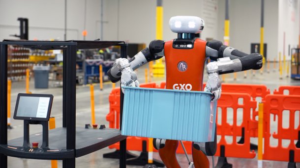 A humanoid robot carries a crate in a warehouse