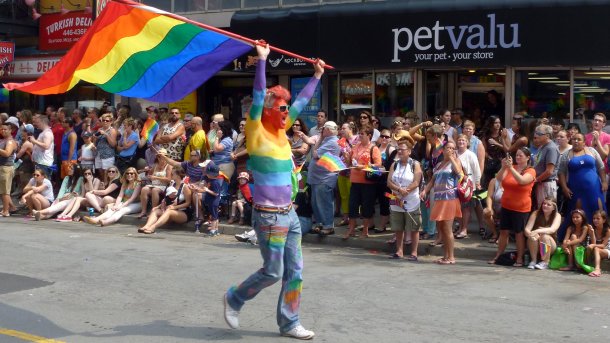 Man with upper body painted in rainbow flags waving a large rainbow flag, with onlookers standing in line behind him