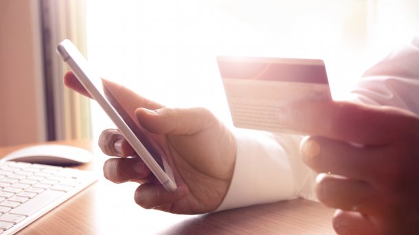 Mensch scheint eine Bezahlfunktion auf seinem Handy zu nutzen. In der rechten Hand hält er n Handy, in der linken eine Kreditkarte.