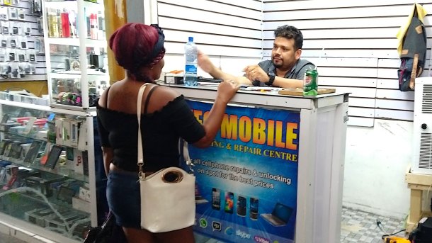Small cell phone repair store in a market hall. A man behind the counter is serving a customer.