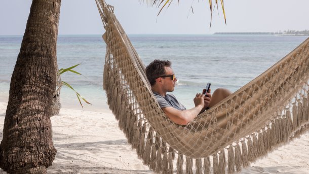Mann kauert im Urlaub auf einer Hängematte am Smartphone und kann nicht abschalten.