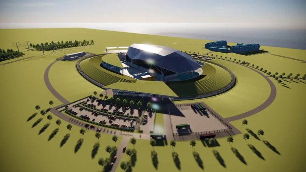 On a green lawn stands a futuristic building with a ring road around it, along which there is a parking lot with a few cars in the foreground; in front of it some young trees; in the background a battery of blue containers, behind it the sea