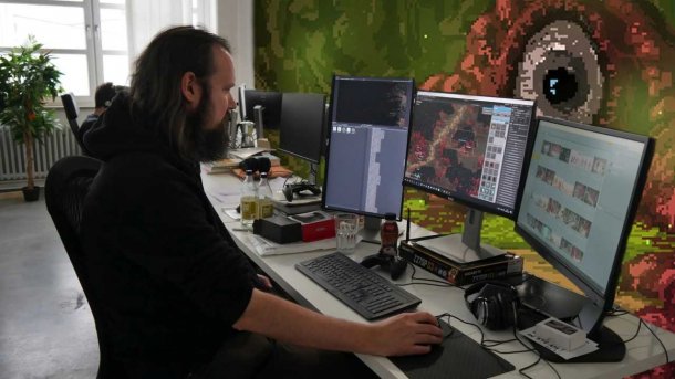 Ein Mann sitzt an einem Tisch und schaut auf einen Monitor, auf dem sein Spiel abgebildet ist. Das Spiel hat eine Pixelgrafik und auch die Wandmalerei hinter den Monitoren hat ein einäugiges Monster abgebildet, das an Cthulhu erinnert.