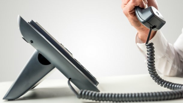 A corded telephone from the side, an arm in a white shirt holds up the receiver.
