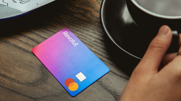 Eine Revolut-Karte liegt auf einem Tisch, daneben Gebäck und eine Tasse Kaffee, nach der eine Hand greift.