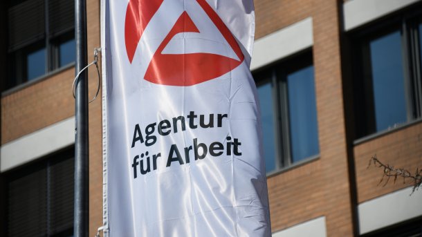 Eine Flagge mit dem Logo der Agentur für Arbeit weht vor einem Bürogebäude.