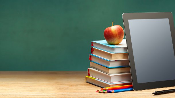 An einen Bücherstapel agelehntes Tablet. Auf dem Bücherstapel ist ein Apfel. Vor dem Tablet liegt ein Stift. Die Wand ist grün, der Tisch, auf dem die Gegenstände platziert sind, aus Holz.