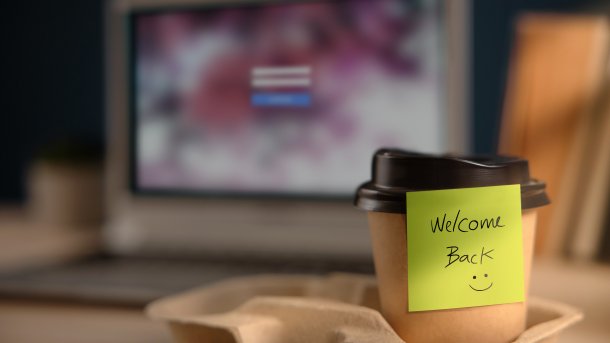 Kaffeebecher mit Zettel "Welcome Back" vor Anmeldebildschirm eines PCs