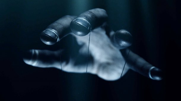 Hand, die eine Marionette steuert. Es ist aber nur die Hand mit den Fäden zu sehen.