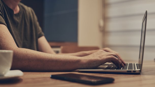 Person nutzt ein Smartphone und sitzt vor einem Computer. Daneben ist ein Stethoskop.