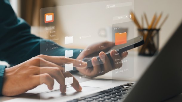 Person vor einem Laptop schaut in ein Smartphone und tippt etwas 