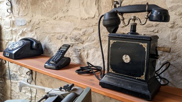 Rechts ein altes Telefon mit Kurbel, in der Mitte ein Schnurlostelefon in Ladeschale, links ein Telefon mit Wählscheibe