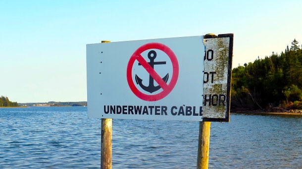 Ein Schild steht auf Holzbeinen im Wasser; es verbietet ankern weil dort ein "underwater cable" verläuft; im Hintergrund grüne Inseln
