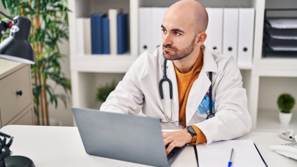 Arzt vor dem Laptop guckt nachdenklich in die Ferne. Um seinen Hals hat er ein Stethoskop. 