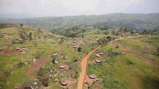 Luftaufanahme einer grünen, hügelischen Landschaft mit wenigen Bäumen und einigen Feldern. Zu sehen sind eine unbefestigte Straße und mehrere niedrige, runde Hütten.
